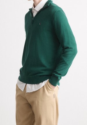 Mann trägt dunkelgrünen Pullover über weißem Hemd mit beiger Hose, Hände in den Taschen, vor schlichtem weißem Hintergrund.