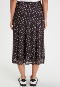 Jupe noire avec motif floral, longueur genou, tissu léger, superposition transparente, taille élastique, design en couches, portée avec des chaussures blanches.