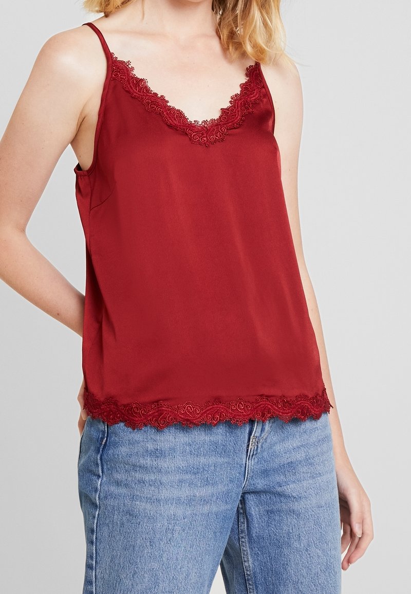 Rød satin top med blondekant ved halsudskæringen og kanten. Spaghetti-stropper og en afslappet pasform, sat sammen med lyseblå jeans.