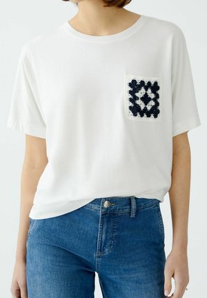 Persoon draagt een wit T-shirt met een zwart-wit gehaakt zakdetail en blauwe spijkerbroek tegen een effen achtergrond.