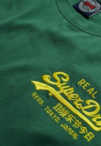 Camiseta de algodón verde con texto bordado amarillo que dice "Superdry" y texto adicional debajo. Textura suave con escote redondeado.