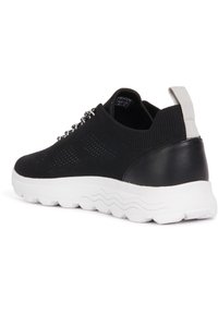 Zapatilla negra con parte superior de punto transpirable, superficie texturizada y talón de cuero negro sólido. Suela de goma blanca que presenta un patrón en forma de ola.