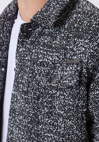 Giacca testurizzata bianca e nera con tasca sul petto con bottone e etichetta metallica del marchio sopra una camicia bianca sul torso superiore di una persona.