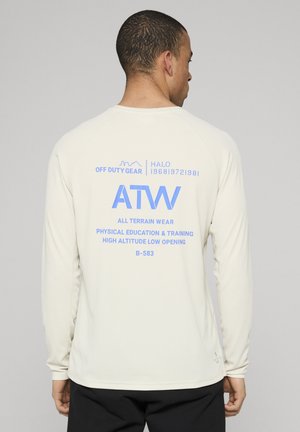 Mężczyzna stojący tyłem, ubrany w kremową koszulę z długim rękawem z niebieskim napisem „ATW All Terrain Wear” oraz powiązanymi hasłami na plecach, w połączeniu z czarnymi spodniami.