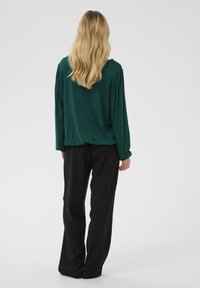 Zeleni top dugih rukava s nabranim ramenima, u paru s crnim hlačama širokih nogavica. Glatki materijal s opuštenim krojem, prikazan s leđa.