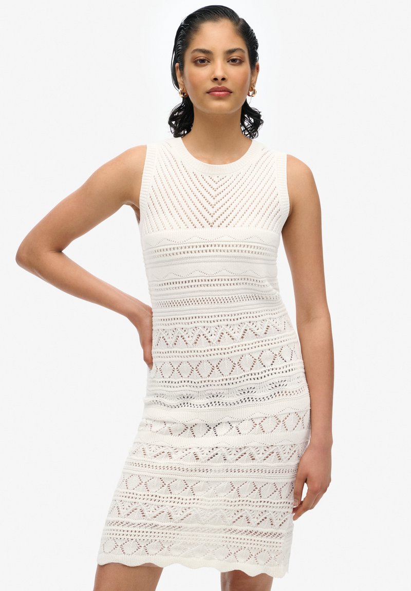 Weißes Strickkleid mit ärmellosem Design, strukturierten Mustern und einer taillierten Silhouette sowie spitzenartigen Verzierungen am Saum.