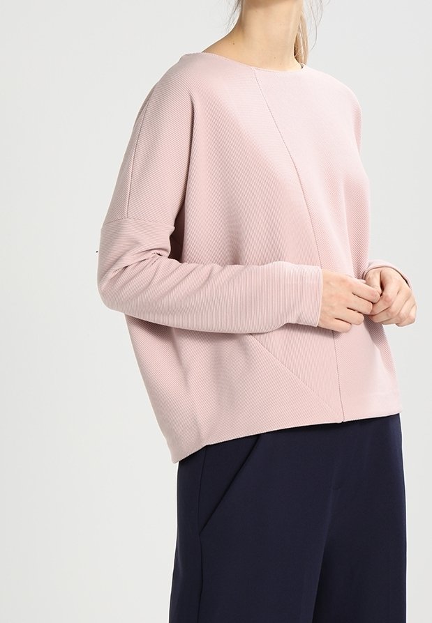 Femme portant un haut ample à manches longues rose clair et un pantalon bleu marine foncé, debout, les mains légèrement jointes à la taille.