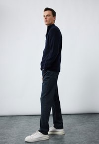 Suéter azul marino de cremallera a medio pecho con tejido texturizado, combinado con jeans de mezclilla oscura y zapatillas blancas. Diseño simple y moderno con líneas limpias.