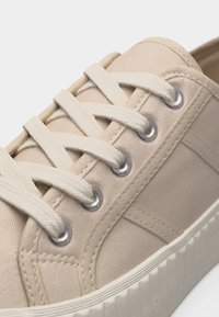 Baskets en toile beige avec semelle en caoutchouc blanche et lacets blancs passant par des œillets métalliques, présentant des détails de couture latérale.