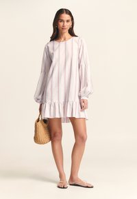 Abito bianco con strisce verticali multicolori, vestibilità ampia, maniche a palloncino, orlo arricciato e una borsa di paglia intrecciata. Si indossano sandali piatti.