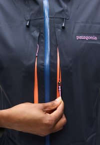 Κοντινό πλάνο χεριού που κλείνει το πορτοκαλί φερμουάρ σε ναυτικό μπλε μπουφάν Patagonia με μπλε κεντρικό φερμουάρ και μοβ κορδόνι έλξης φερμουάρ.