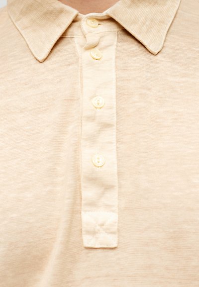 Μπεζ πλεκτό πόλο πουκάμισο με υφή υφάσματος, κλασικό γιακά και πέντε λευκά κουμπιά κατά μήκος της πατιλέτας. Διακριτική διαφορά στο χρώμα του υφάσματος.