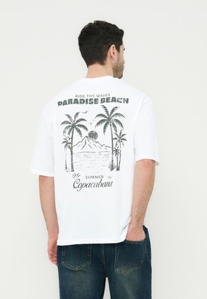 Άνδρας με λευκό μπλουζάκι που έχει γραφικό με φοίνικες, πουλιά, βουνό και ωκεανό, με κείμενο στο πίσω μέρος που λέει «Paradise Beach, Summer Copacabana».