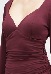 Maroon long-sleeve top featuring a deep V-neck, fitted bodice with ruching on the sides, and smooth, stretchy fabric texture.