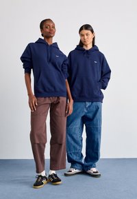 Hoodies azuis marinhos com bolsos frontais e cordões, combinados com calças largas castanhas e azuis. Ténis pretos com sola de borracha e meias estampadas.