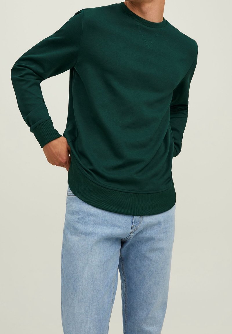 Homme portant un sweat-shirt vert foncé à manches longues et un jean bleu clair sur un fond uni clair, mains près de la taille.