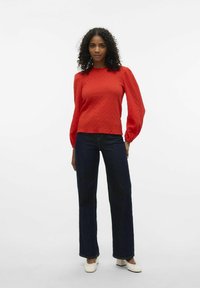 Rote, strukturierte Bluse mit Puffärmeln, kombiniert mit dunkelblauen, weit geschnittenen Jeans und weißen Blockabsatzschuhen. Schlichtes, modernes Design.