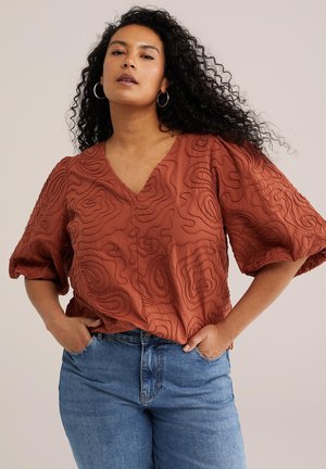 Frau mit lockigem Haar, die eine rostfarbene Bluse mit Puffärmeln und Textur sowie eine blaue Jeans trägt, mit den Händen in den Taschen stehend.