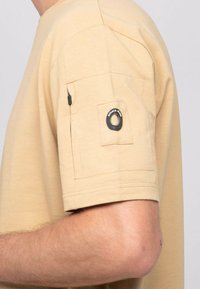 Chemise beige à manches courtes en tissu texturé, dotée d'une poche sur la manche et d'un accent en métal noir sur la poche.