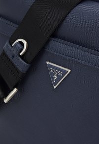 Borsa in tessuto testurizzato blu navy con patch logo a forma di triangolo argentato, tracolla nera con hardware in metallo e dettagli cuciti.