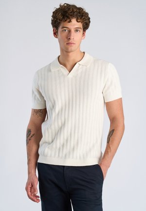 Junger Mann mit lockigem Haar, der ein weißes, kurzärmeliges, geripptes Poloshirt und dunkle Hosen trägt, steht mit einer Hand in der Hosentasche.