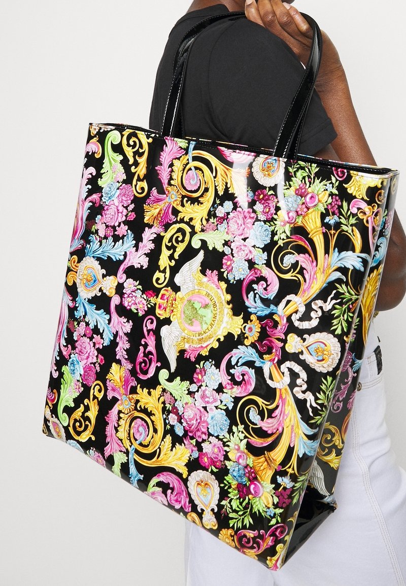 Personne tenant un grand sac cabas noir brillant avec un motif floral et orné coloré à volutes, avec de courtes poignées noires.