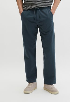 Mann trägt dunkelblaue Hosen mit Kordelzug und hellbeige Sneaker, Hände in den Taschen, steht vor einem schlichten hellen Hintergrund.