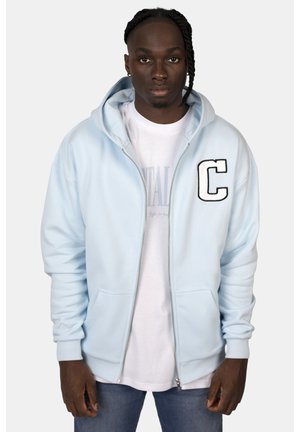 Hellblaues Kapuzenshirt mit Reißverschluss, einer vorderen Tasche und einem großen weißen 'C'-Patch. getragen über einem weißen T-Shirt und kombiniert mit blauen Jeans.