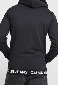 Osoba v černé mikině s kapucí a nápisem "Calvin Klein Jeans" na pase, vyobrazená zezadu na neutrálním pozadí.