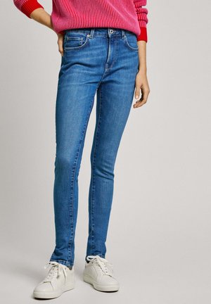 Dunne blauwe spijkerbroek van denim, met een klassiek vijf-pocket ontwerp, een glanzende metalen knoop en subtiele stiksel details. Gecombineerd met witte sneakers.
