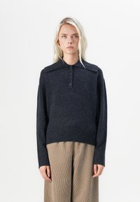 Ciemnoszary sweter z dzianiny z szerokim kołnierzem, zapinany na trzy guziki, z prążkowanym dołem i mankietami, noszony na tle wzorzystych brązowych spodni.