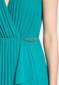 Robe plissée en teal avec un design enveloppant, offrant un détail de ceinture en chaîne dorée. La texture douce et le tissu fluide améliorent la silhouette.