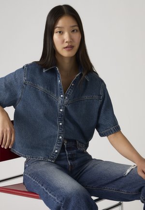 Młoda kobieta z długimi ciemnymi włosami siedząca na czerwonym krześle, ubrana w luźną jeansową koszulę i dopasowane podarte jeansy na jednolitym tle.