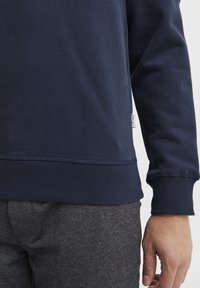Marinblå sweatshirt med ribbade ärmslut och nederkant. Tillverkad av strukturerad tyg och har en liten etikett på sidan. Kombinerad med grå strukturerade byxor.