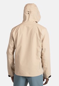 Beige waterdichte jacket met een capuchon, met een gladde textuur, lange mouwen en een logo-detail op de achterkant. Ontworpen voor buitengebruik.