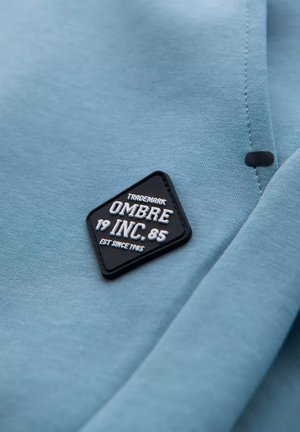 Světle modrá tkanina s texturovanou černou diamantovou nášivkou, na které je bílé písmo a čísla: "OMB 19 INC. 85 EST SINCE 1985."