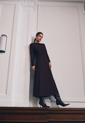 Femme portant une longue robe noire à pois et des bottines noires à talons, appuyée contre un mur blanc avec une moulure décorative.