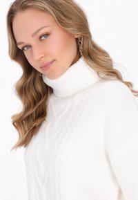 Weißer Strickpullover mit hohem Rippkragen und Zopfmuster. Weicher Stoff, lockere Passform und mit leicht abfallenden Schultern.