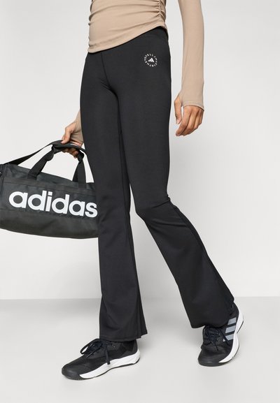 Μαύροι κολάν με φαρδύ κάτω μέρος και λεπτομέρεια με λογότυπο στο ισχίο. Συνοδεύονται από μαύρη αθλητική τσάντα με το εμφανές λογότυπο "adidas" σε λευκό και ένα ζευγάρι μαύρα αθλητικά παπούτσια.