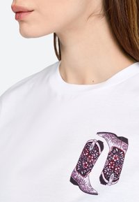 T-shirt blanc avec un design imprimé de deux bottes de cowboy décoratives en violet et rouge avec des motifs floraux et des embellissements.