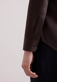 Donkerbruin overhemd met lange mouwen en een gladde stofafwerking. Voorzien van een knoopmanchet, een lichte glans en een getailleerd ontwerp. Met een schaduwachtige achtergrond.