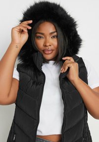 Musta puffertakki, jossa on tekoturkis-huppu, varustettu vetoketjulla ja tikatulla pinnalla. Mallilla on päällään valkoinen yläosa ja hän poseeraa kädet hupun lähellä.