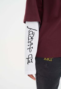 Bordowa koszulka z krótkim rękawem nałożona na białą koszulę z długim rękawem, z czarnym tekstem graficznym. Czarne spodnie z żółtym haftem.