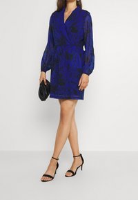 Blå wrapklänning med ett svart blommönster, långa puffärmar och en smal midja; ihop med svarta remsandaler och en clutch.