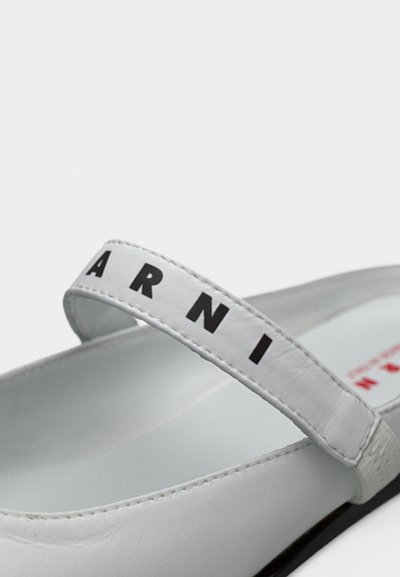 Λευκά επίπεδα σανδάλια με λουράκι που φέρει μαύρο λογότυπο. Το υλικό είναι λείο και γυαλιστερό, τονίζοντας ένα μινιμαλιστικό σχέδιο.