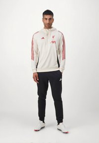 Vit luvtröja med röd rand och Liverpool FC-logotyp, kombinerad med svarta joggerbyxor och vita sportskor.