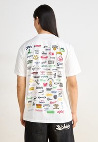 Camiseta blanca con varios logotipos de marcas coloridos impresos en la parte posterior. Diseño de mangas cortas y cuello redondo. Pantalones negros con detalle del logotipo.