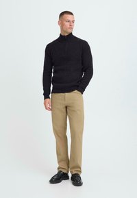 Schwarzer, langärmliger Pullover mit hohem Kragen und einem halben Reißverschluss, kombiniert mit beigen Hosen und schwarzen Schuhen. Einfaches Design und strukturierter Stoff.