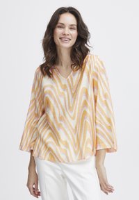 Chiffon bluse med V-udskæring, der har et b�ølget mønster i orange, gul og pink. Flared ærmer og en løs pasform for komfort. Hvide bukser bæres nedenunder.