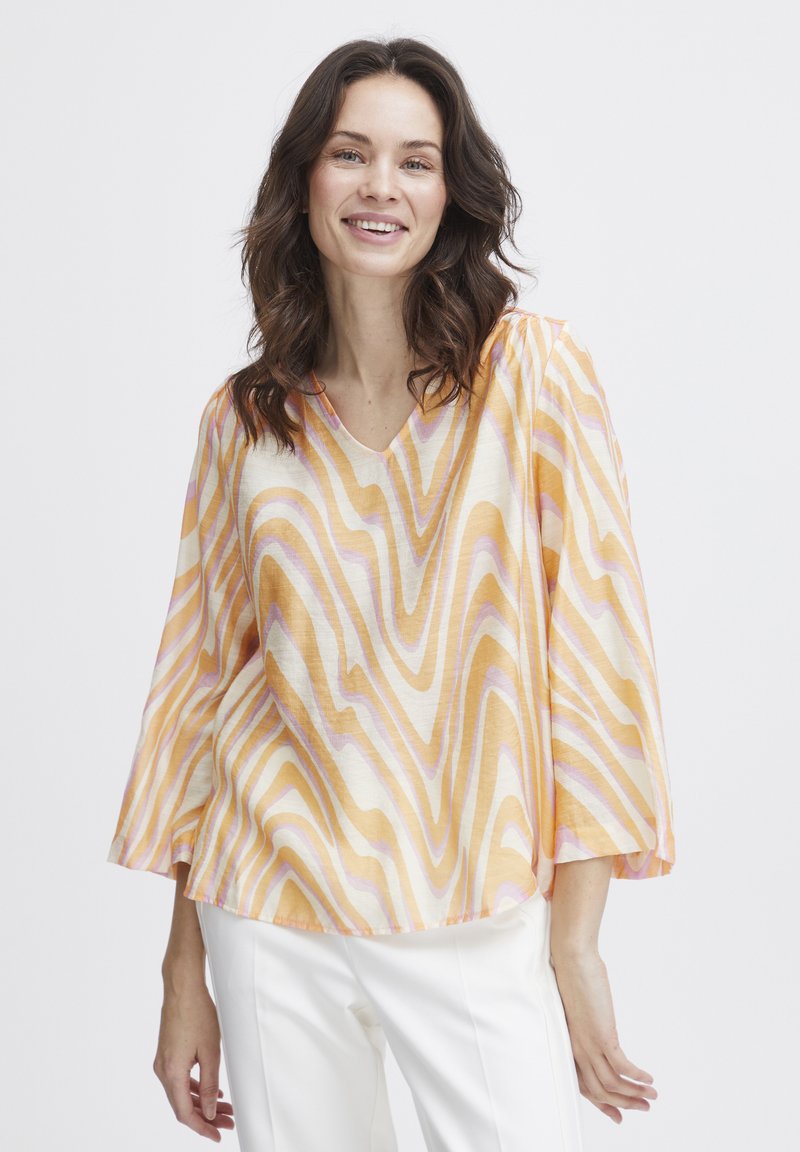 Chiffon bluse med V-udskæring, der har et bølget mønster i orange, gul og pink. Flared ærmer og en løs pasform for komfort. Hvide bukser bæres nedenunder.